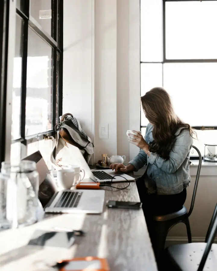 A person with a cup of tea working on a laptop at a desk by a bright window